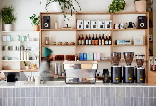 Behind the bar at Prince Coffee. Wide open shelves display cups, syrups, bags, and equipment. The bar has vertical light blue thin tiles. Plants hang overhead a La Marzocco silver machine, with grinders to the right.