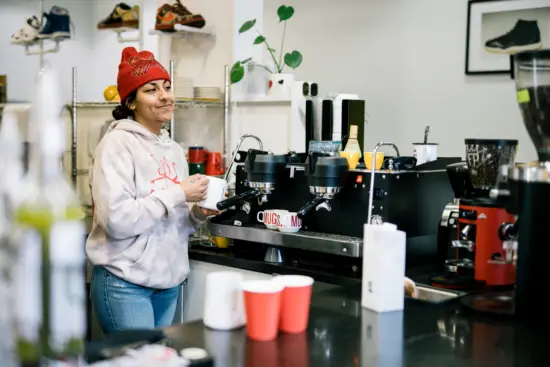 A barista in a red beanie and white hoodie steams milk at the espresso machine behind the bar at Deadstock. Sneakers and a plant are displayed on the wall behind the bar.