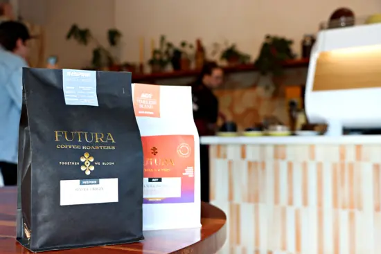 Two coffee bags from FUTURA, one black and one white. The print is too small to read except for the logo: FUTUTRA COFFEE ROASTERS: together we bloom. The shop espresso machine is blurred in the background.