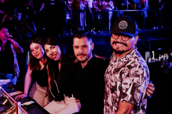 Four people pose for the camera by the bar. Two women have long brown hair, two men have mustaches; one wears glasses and a hat.