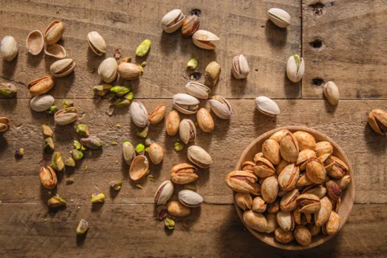 A small wooden bowl of whole pistachios, with cracked shells and nuts scattered around on a wooden table.