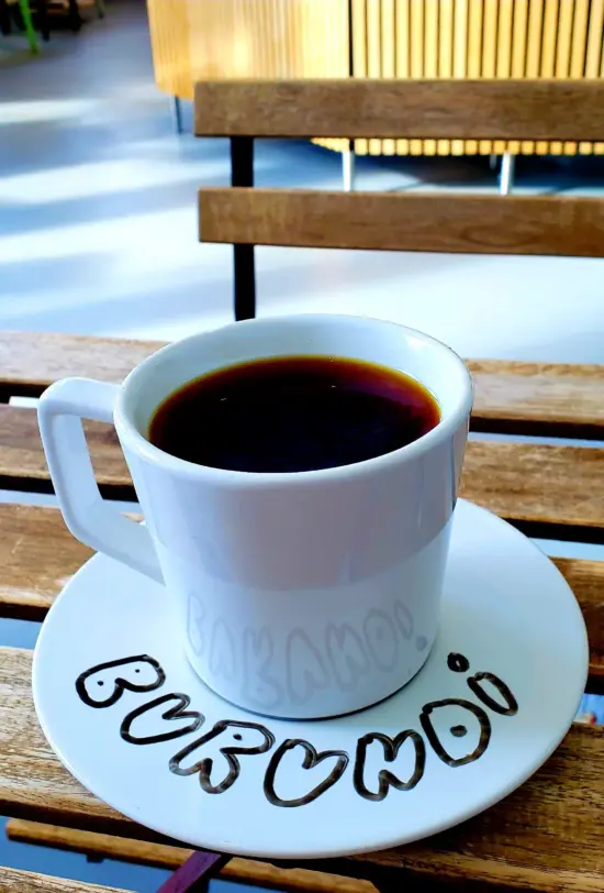 A plain white mug of coffee on a wooden outdoor table, on a white saucer with the Word Burundi written on it in bubble letters.