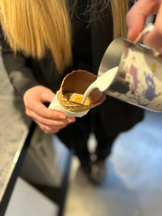 A barista with long blonde hair pours milk into a waffle cone.
