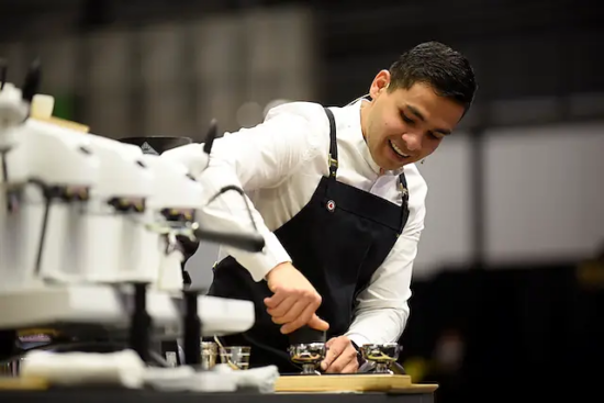 Diego wears a white shirt, black apron and is tamping an espresso shot while speaking into his headset during competition.
