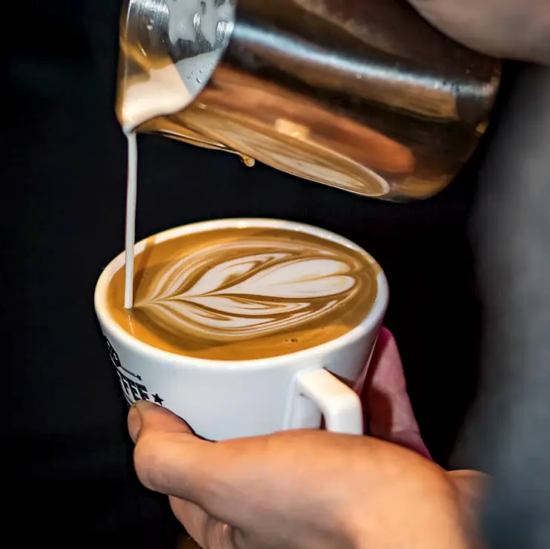 A close up of a barista pouring milk from a metal pitcher into a cappuccino cup, making a tulip.