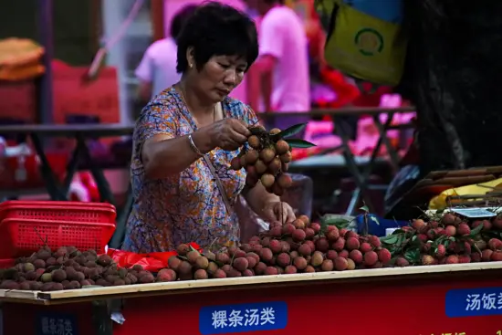 A woman in China looks over lychee bundles in a big market stall, holding up a big cluster. To her right is a stack of red plastic hand baskets, and people browsing are visible behind her.