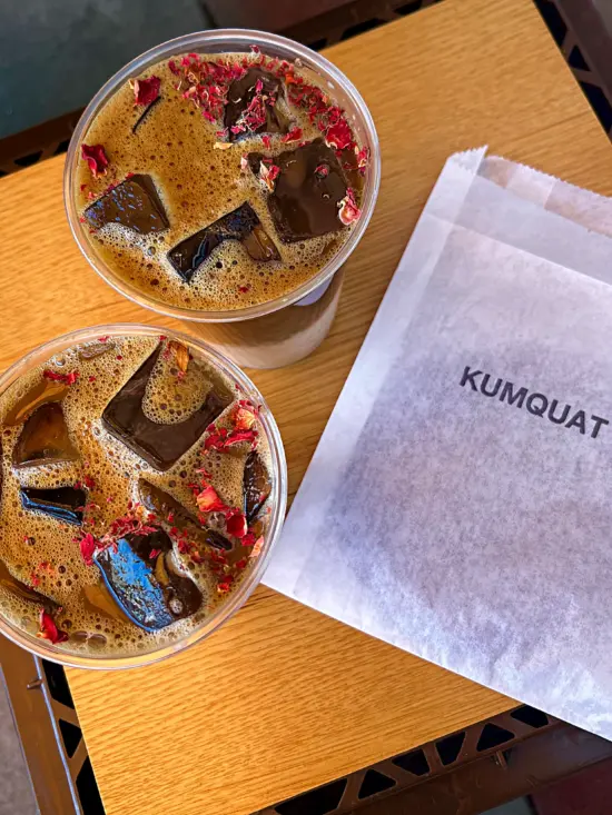 A white paper bag labeled KUMQUAT and two lychee iced tea drinks with rose petal pieces sprinkled on top. The drinks are layered so milk sits at the bottom.