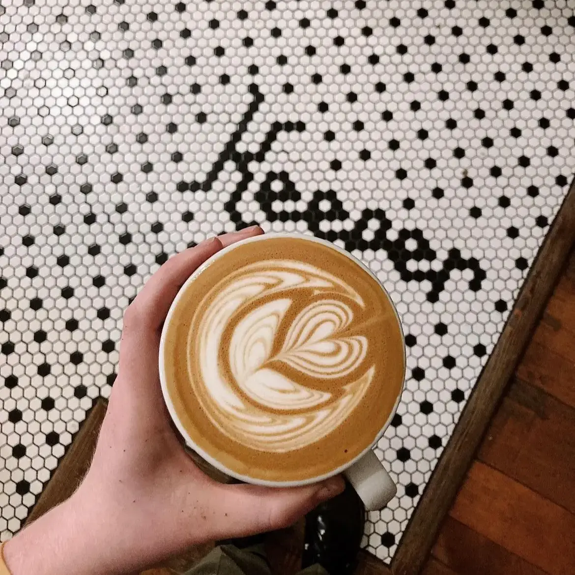 A hand holds a mug with a latte featuring a rosetta. The countertop underneath is covered in tiny hexagon tiles, spelling out Keeper in tile right above the drink in the photo.