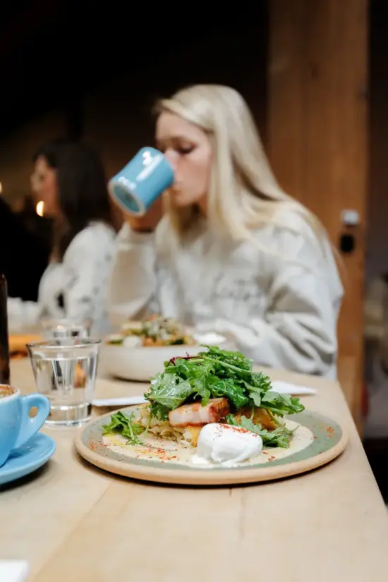 A customer sips a drink behind a beautiful plate of poached eggs and greens with meat.