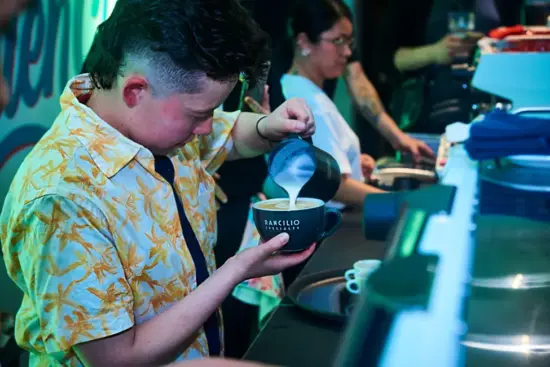 Carolina wears a yellow flowered shirt and pours a latte into a big mug. Behind her other baristas are also working the bar.