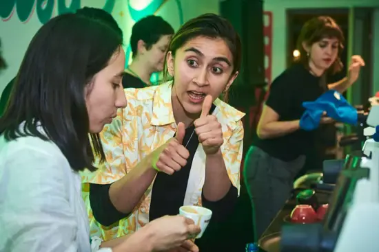 Lorena communicates to Ivette with thumbs up as they taste coffee from a small cappuccino cup.