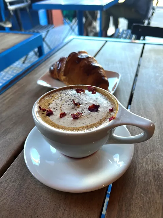 A pistachio latte with rose petals and cardamom powder sprinkled on top. The latte is in a white notNeutral mug with a large handle and rests on a white saucer. It is on top of a wooden cafe table, with a croissant on a plate behind it and a blurred view of the rest of the seating area.