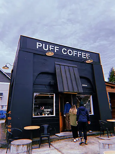 Outside Puff, the building is painted black with PUFF COFFEE in huge white lettering at the top. Two small square windows decorate the front, ans a wooden slat hangs above the entrance, also painted black. Small cafe tables are placed out front.
