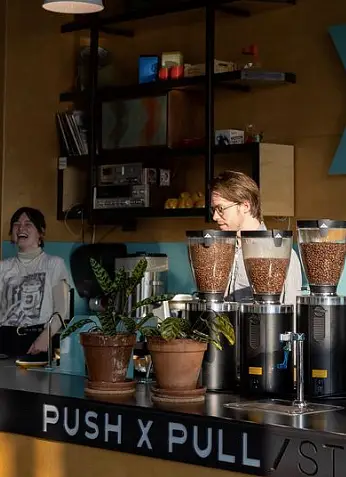 The bar at Push X Pull. Two baristas talk and laugh as they work, while three grinders and two potted plants sit on top of the bar. The wall behind is painted blue and brown and has an open shelf.