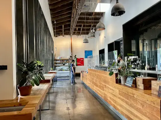 Inside the first location are potted plants and a long wooden bar running the length of the shop. Industrial lamps and wooden exposed beams adorn the ceiling. 
One wall features enormous windows.
