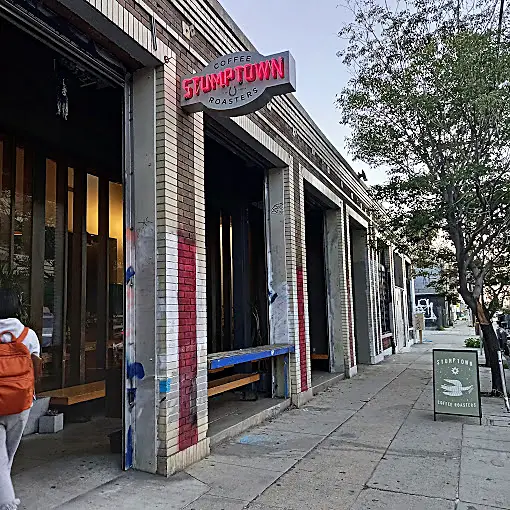 The brick extorior of Stumptown, with wooden benches on the covered porch, a sign with a dove out front on the sidewalk, and a neon red sign sticking out above the building.