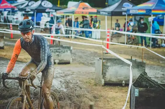 Andrew takes a corner of the dirt track in a bike competition. He's on a muddy bike, wearing blue and orange with a black helmet, all spattered in mud. 