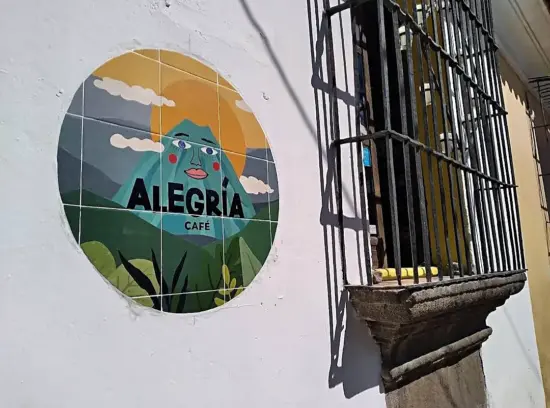 The barred window outside Alegria, on a white wall, with a sun and mountain logo painted next to it.