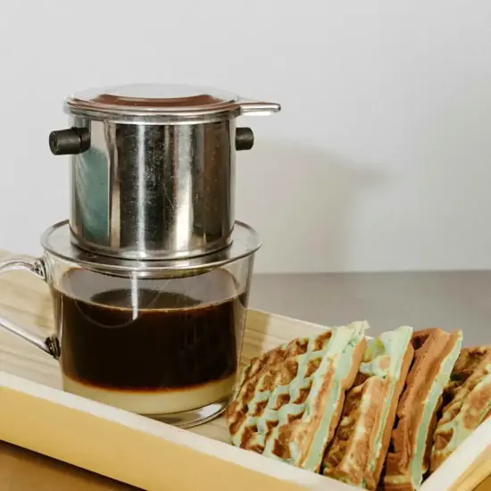 A silver phin filter over a clear glass mug with coffee brewed into it, over a layer of condensed milk on the bottom. It sits on a small tray with a sliced pandan waffle in a traditional waffle shape.