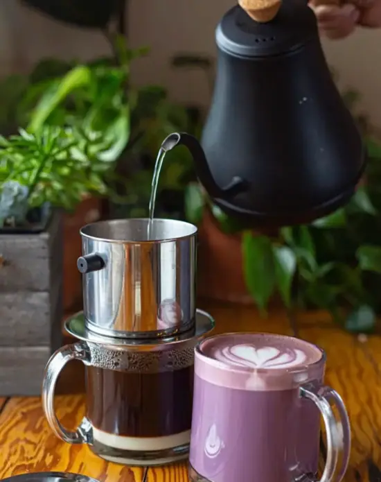 Water is poured from a gooseneck kettle over a phin filter. The filter sits on a clear glass mug and has condensed milk in the bottom. On the table beside it is a clear glass mug with a purple ube latte, which has a rosetta design poured into it. There are potted plants on the table behind the drinks.