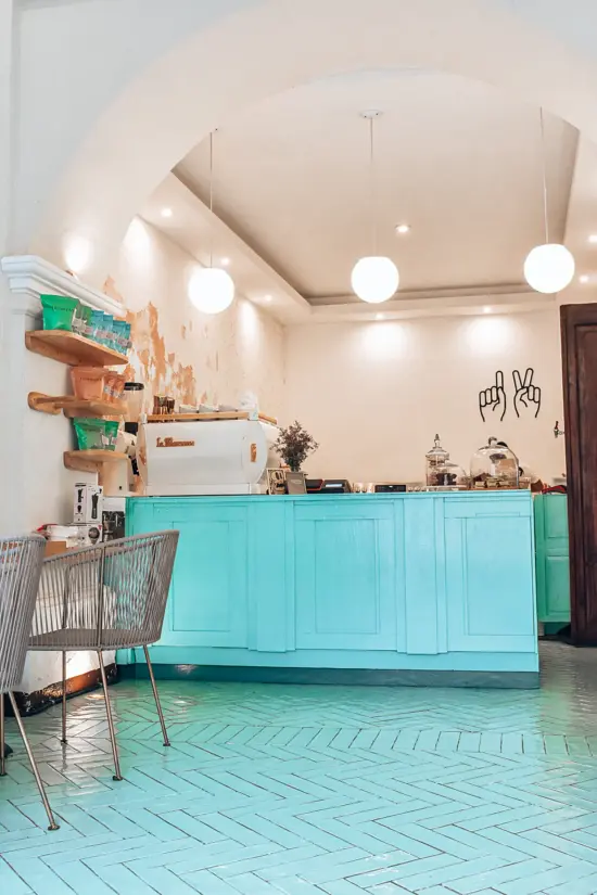 Inside 12 ONZAS, the floor is zigzag blue tiles, with a matching blue bar, white circle pendant lights under an arch, wire chairs and white walls and espresso machine.