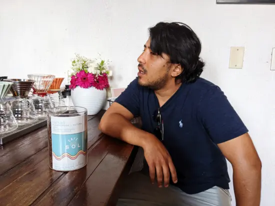 Felipe sits at a wooden counter at Cafe Sol, which brews his coffees. V60s line the bar and there is a white vase with pink and white flowers at the end.