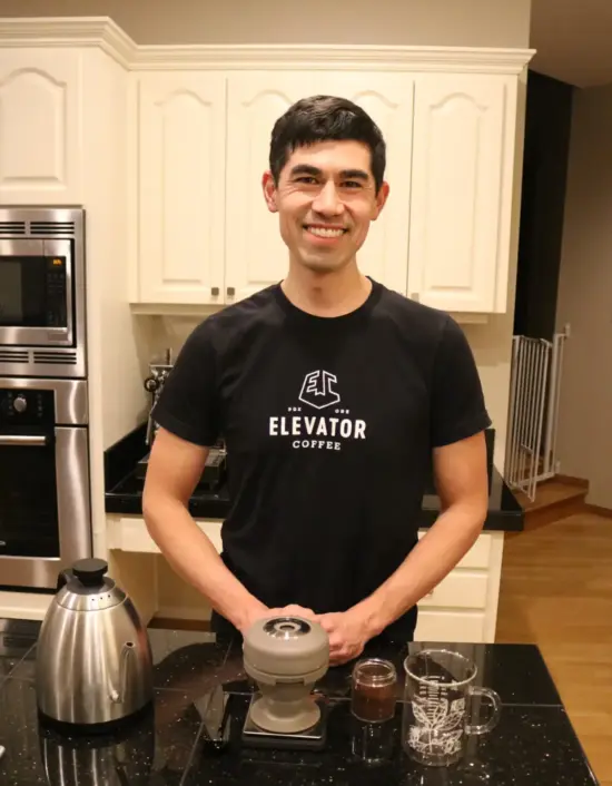 Andrew in his kitchen, which has a black marble counter, and a coffee setup for brewing. He wears a black Elevator Coffee Logo shirt and smiles.