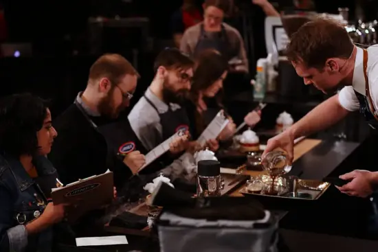 Isaiah is pouring drinks for the judges at a dark bar in the 2018 semifinals. Judges wear black aprons and score on their clipboards. Isaiah is in an apron and white button up shirt and tie.