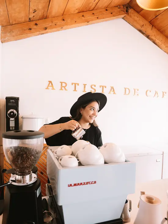 Maria pours a latte behind her white La Marzocco. She wears a large black hat. On the white wall behind her, the cafe name is spelled out in all caps.