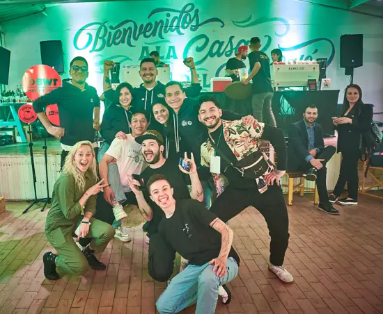 A group of people  make silly poses for the camera at a Barista League event. Juan is in the middle, on his knees and making devil horns with his hands.