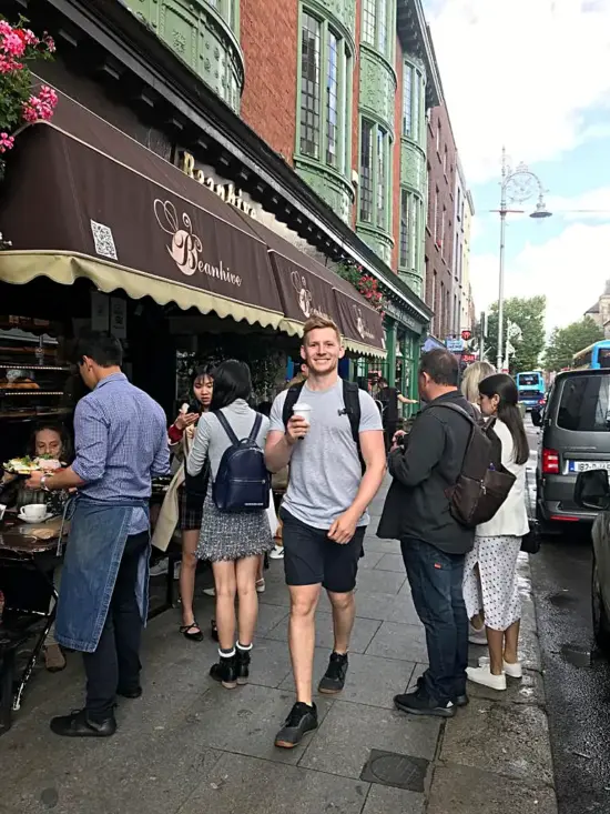 Mitchell, a tall redheaded man, wears a backpack and walks down an Irish street holding a coffee to-go cup.