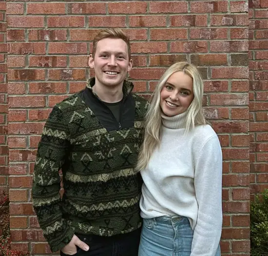 Mitchell on the left wears a knit sweater and Bethany, with long blonde hair, wears a white turtleneck and jeans. They stand in front of a red brick wall. 