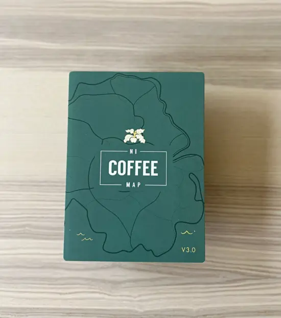 A wood grain table top with the dark green NI Coffee Map. The map is folded closed, with illustrated map lines in black on the green background, and a white flower above a white box outline with the word coffee in bold, an NI written above it inside the box line, and Map written below inside the box line.