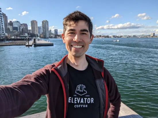 Andrew takes a selfie by blue water in front of a city skyline. Fluffy clouds dot the sky.