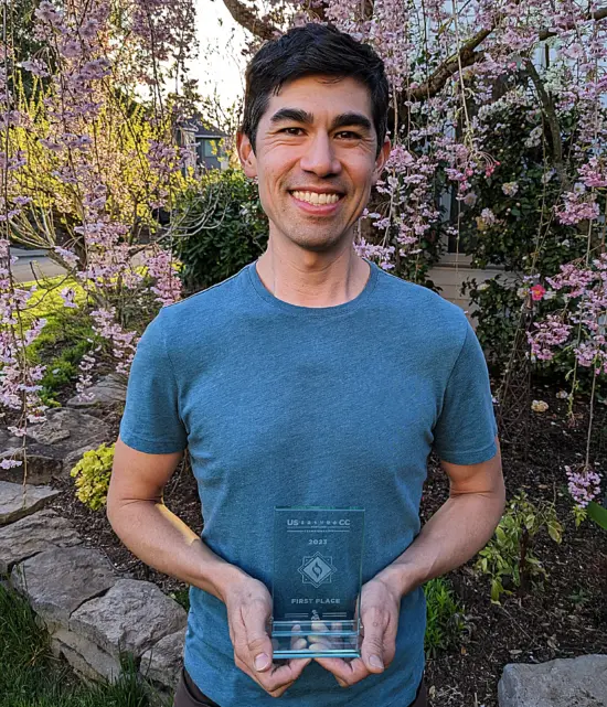 Andrew stands in a garden in front of a tree cascading pink flowers on its branches. He holds up his first place glass trophy for the US Coffee Champs Roasting Championship. He wears a blue tee shirt.