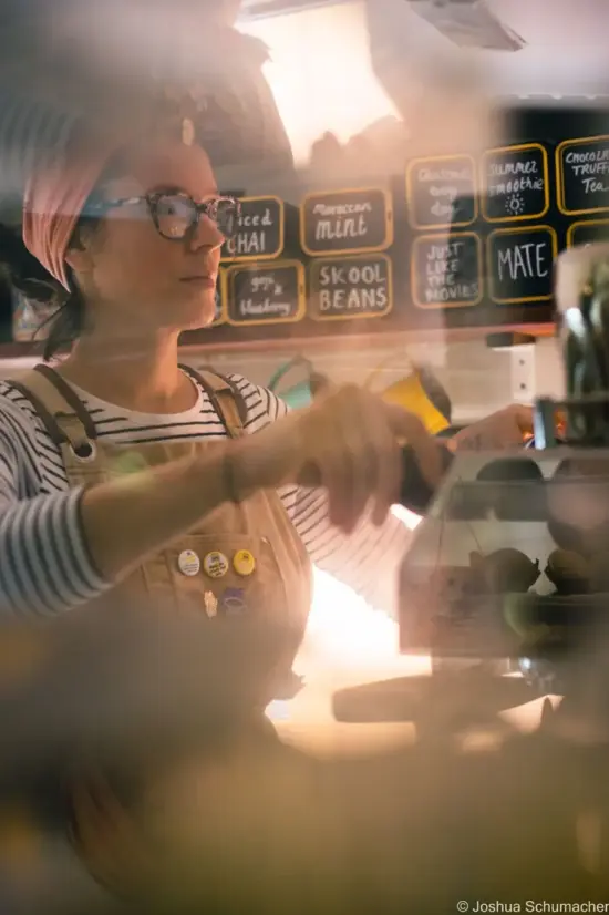 Holly making a drink inside the bus, seen through the window. She wears a headband and khaki apron and has her hands placed on knobs on the espresso machine.