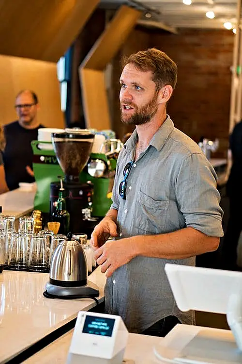 Isaiah at Archetype Coffee in 2018. He stands at the bar in front of a ketter and glassware. Behind him is a green espresso machine.