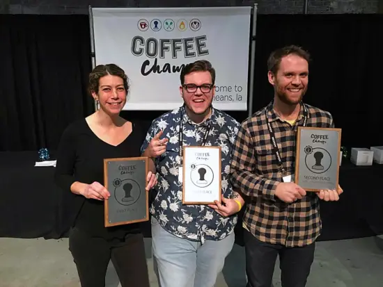 Isaiah, on the right, holds up a wooden plaque with a picture of a tamper and Coffee Champs logo, that reads Second Place. He wears a flannel shirt and lanyard. In the middle is a floral shirt-and glasses-wearing Jared Jolt and Andrea Allen in all black.