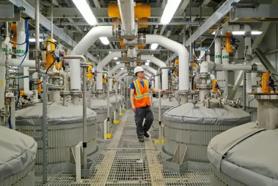 An engineer in hardhat and safety vest looks over the columns above enormous coffee tanks in the facility. The white tubes lead from the ceiling to the tanks, which are grey and low to the floor. 