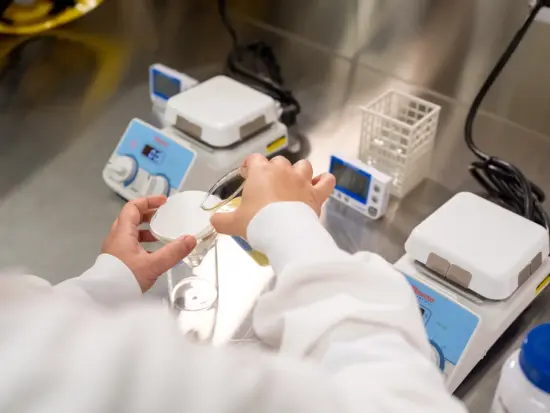 A chemist in a lab pours liquid into a glass vial for testing at the Swiss Water facility. On the table are two scales timers, and a bottle with blue lid. 