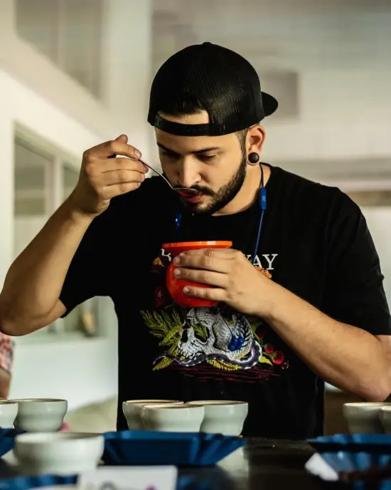 Juan slurps coffee from a cupping spoon at Nordic Approach. He wears a black tee shirt and backwards black ballcap.