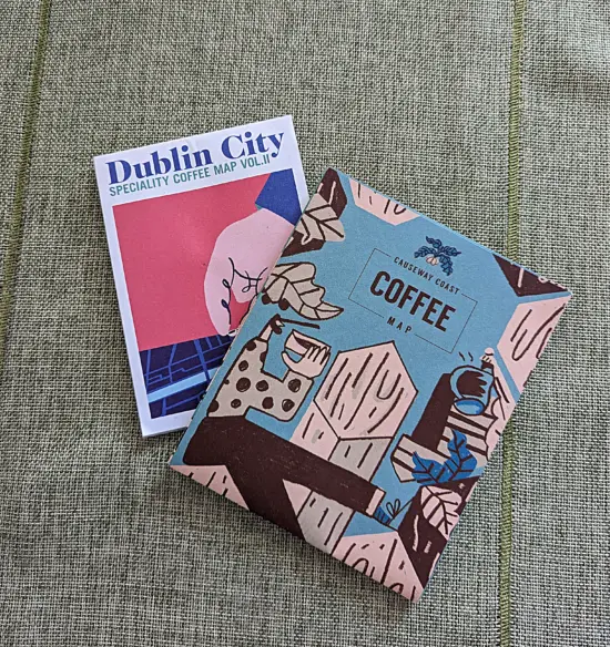 Two coffee maps. On the left is one for Dublin City. On its cover is an illustrated hand on a bright pink background, pointing to a spot on a blue map surface. On the right is the Causeway Coast map, which is blue and has black and white illustrations of people with tiny heads and exaggerated bodies drinking coffee out of mugs.