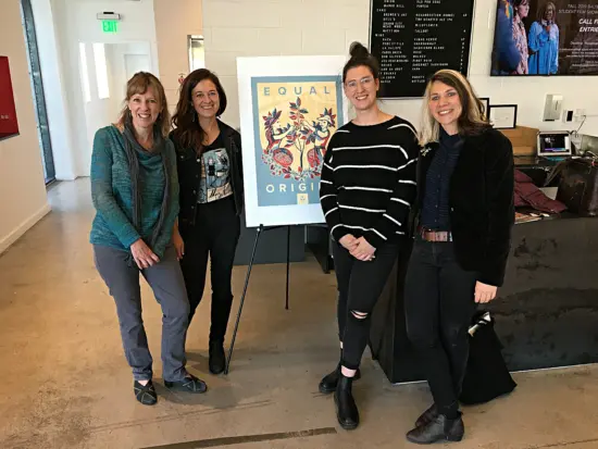 Four women stand together in a coffee shop in front of a sign that reads "Equal Origins." 