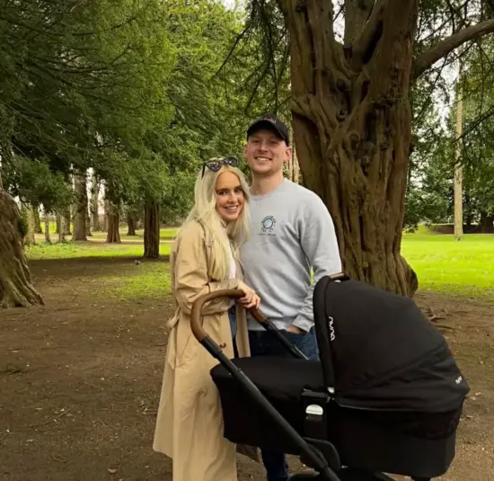Bethany and Mitchell pose in front of a big tree in a park with green moss on the ground. They are pushing a black stroller. 