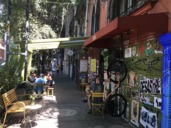 The outside of Cumbé has a green garage door with a mounted bike rack, is covered in stickers, has a green patio covering, and yellow seating.