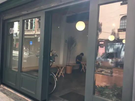 The gray doorframe and windows outside Curva. Inside you can see a potted tree, small tables and a long bench, with a bicycle leaning against the wall by the door. To the right is a silver espresso machine and plant in front of it.