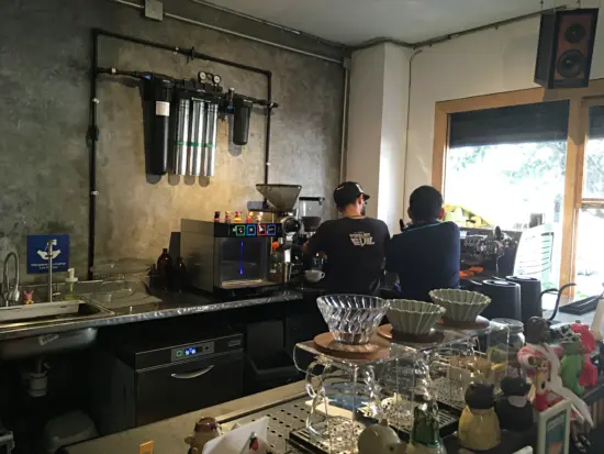 Behind the bar at Fuzz and Brew, two baristas work in the corner by the espresso machine. There are Origami and V60 pourover setups and Fellow kettles on the bar, along with small cartoonish figurines.