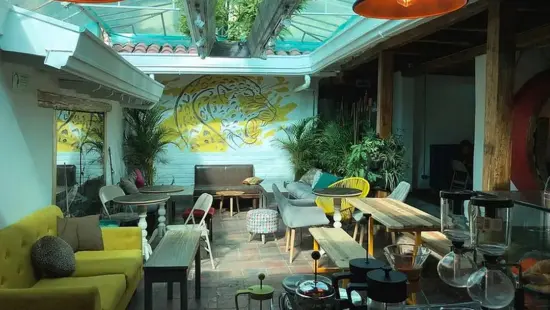 Inside Jaguar's cafe space, bright blue and yellow walls surround the seating area. A glass roof allows sunlight to filter in. Palm trees are set up in the corners. Seating includes a lime green couch with gray pillows, small wooden tables, and a variety of gray  and yellow chairs and ottoman.