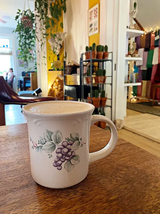 A white porcelain mug painted with a bunch of grapes inside a cafe, on a wooden table.