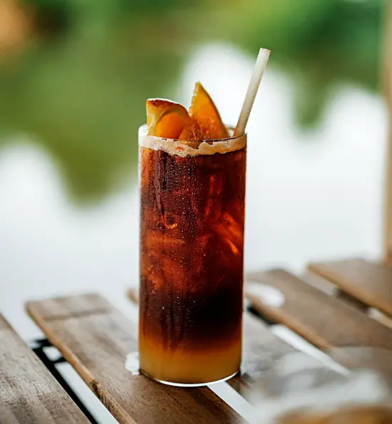 a tall skinny cylinder glass with a brown cocktail and orange garnish and white straw. It sits on a slatted wooden table outside, and the background is blurred.